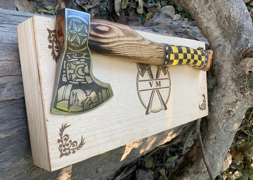 Fenrir Viking Axe with decorative handle and blade, placed on a wooden box with engraved designs, against a natural background.