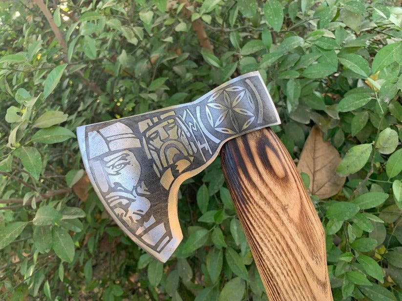Decorative Fenrir Viking Axe with engraved design on a wooden handle against a green leafy background