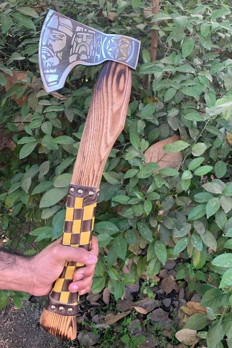 Hand holding a decorative Fenrir Viking Axe with a checkered handle against a green leafy background