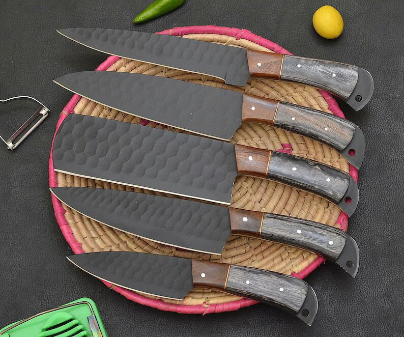 A set of five chef knives with black powder-coated stainless steel blades and ergonomic Rosewood & Wenge wood handles, displayed on a red textured surface, with a leather sheath beside them.
