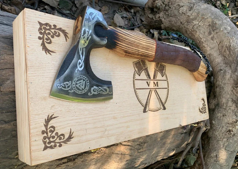 Valknut Viking Axe with wooden handle and black blade on a wooden box with engraved designs, outdoors.