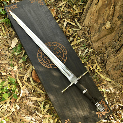 A Viking-style sword with a silver blade and black handle lies diagonally on a dark wooden box printed with Norse runes, set outdoors on dry leaves and tree bark.