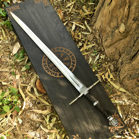 A Viking-style sword with a silver blade and black handle lies diagonally on a dark wooden box printed with Norse runes, set outdoors on dry leaves and tree bark.