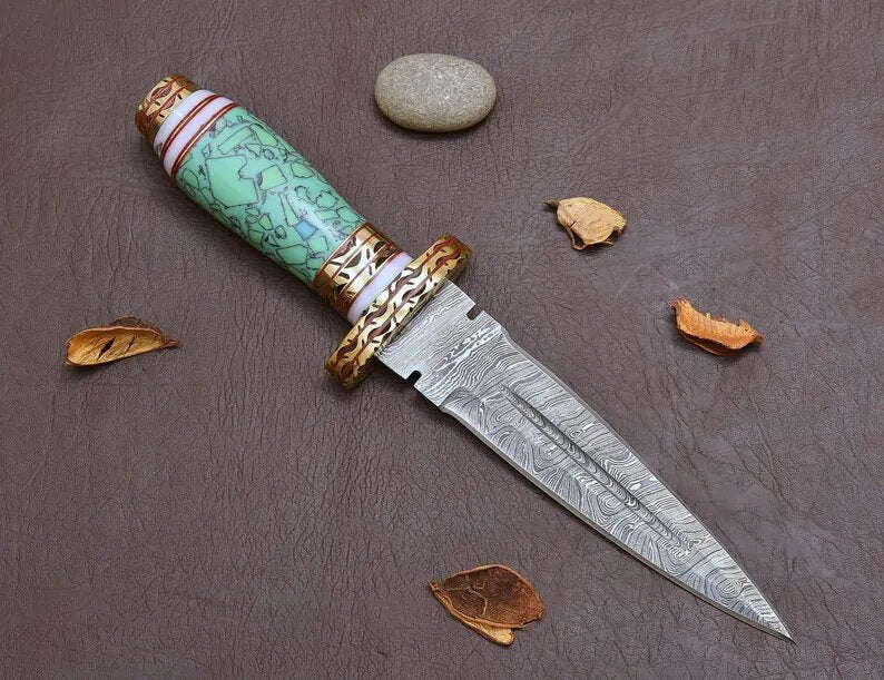 Decorative dagger with a blue resin  handle and gold accents on a brown surface with leaves and stones.