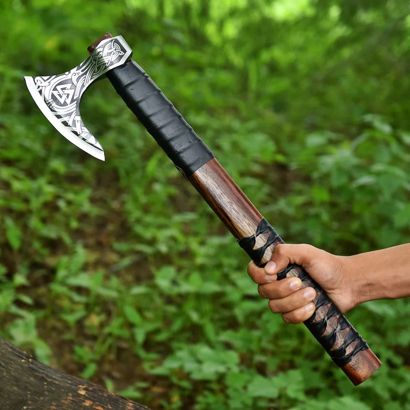 Hand holding a decorative axe with a wooden handle against a green forest background
